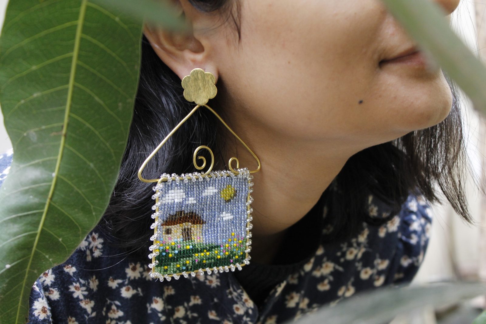 A small beaded earring shaped like a framed cottage scene, hanging from a wire hanger