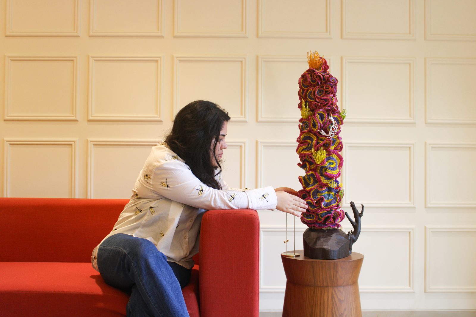 Yashica adjusting a tall, densely beaded coral-coloured sculpture standing on a wooden base beside a red sofa